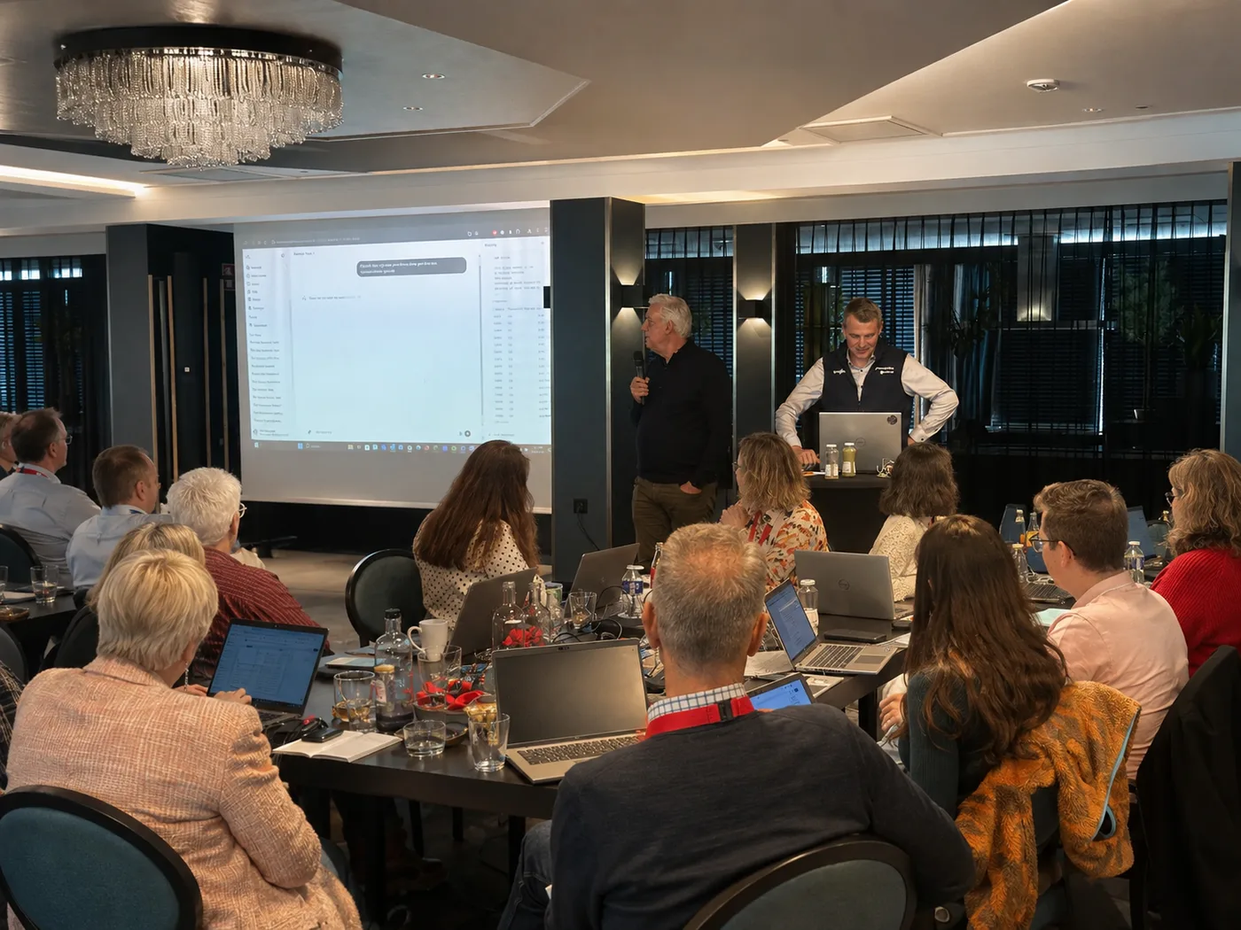 Twee trainers geven een live demo op het grote scherm, de zaal kijkt mee met de laptop open.